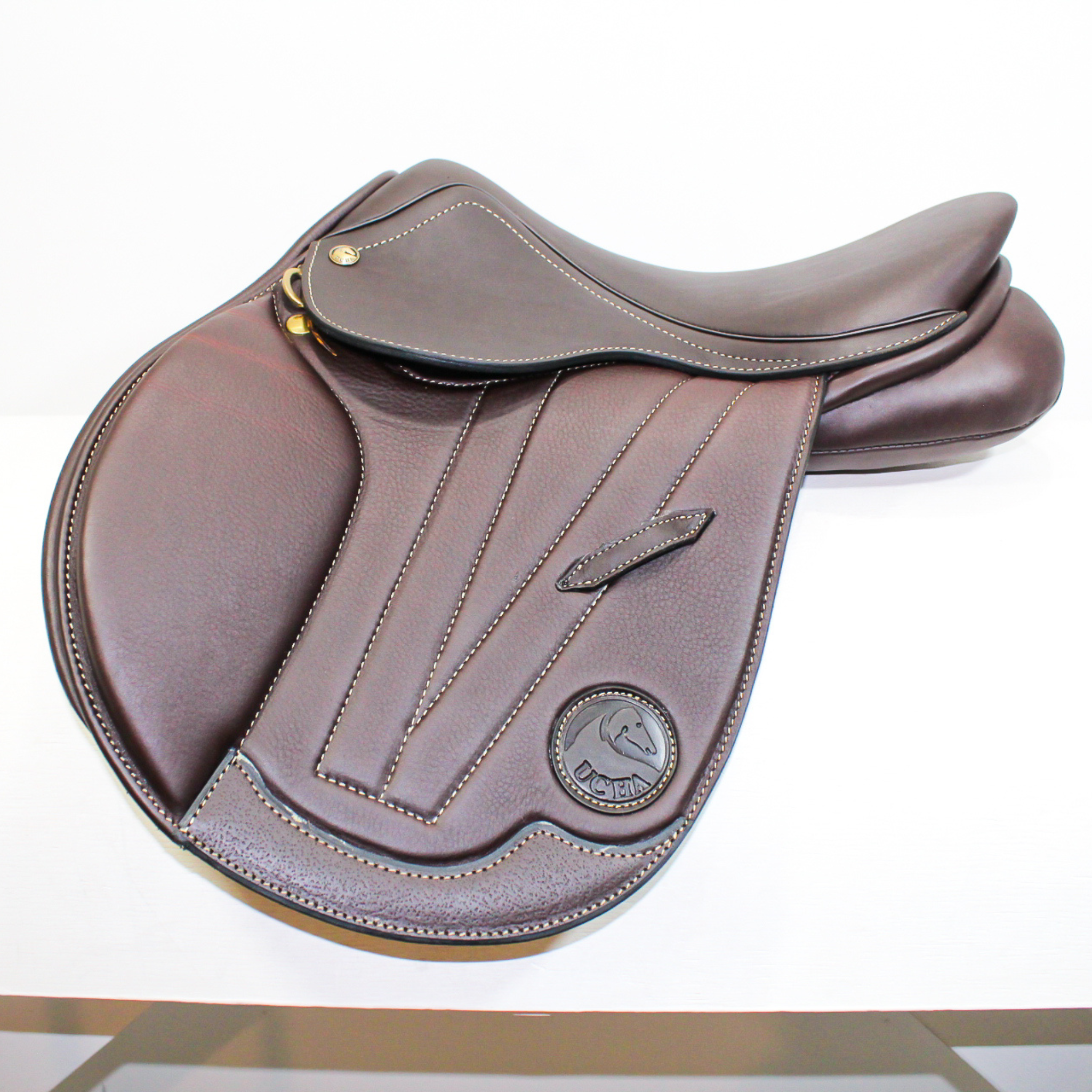 Left side profile view of an Ucha Joven dual flap jump saddle in dark brown calf leather, with a leather Ucha logo and decorative stitching on the flap.