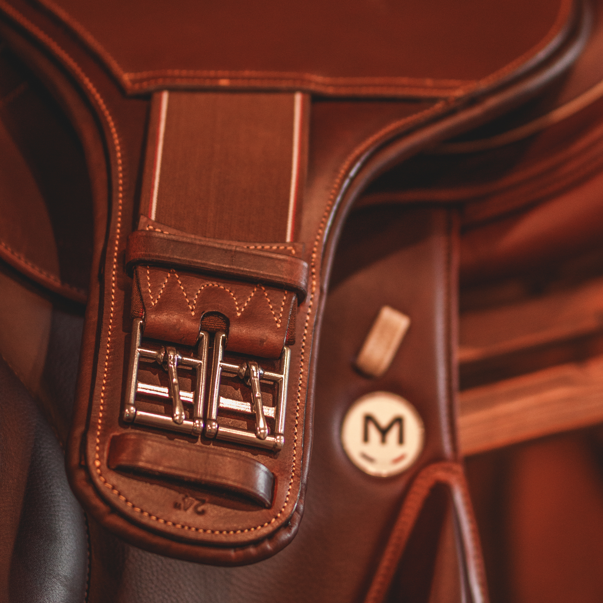 A brown leather short stud girth draped over a Meyer monoflap saddle. The photo focuses on the girth buckles.