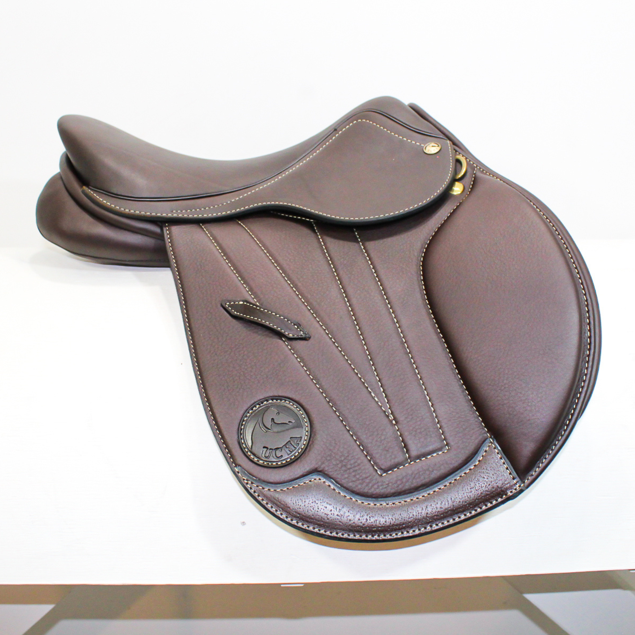 Right side profile view of an Ucha Joven dual flap jump saddle in dark brown calf leather, with a leather Ucha logo and decorative stitching on the flap.