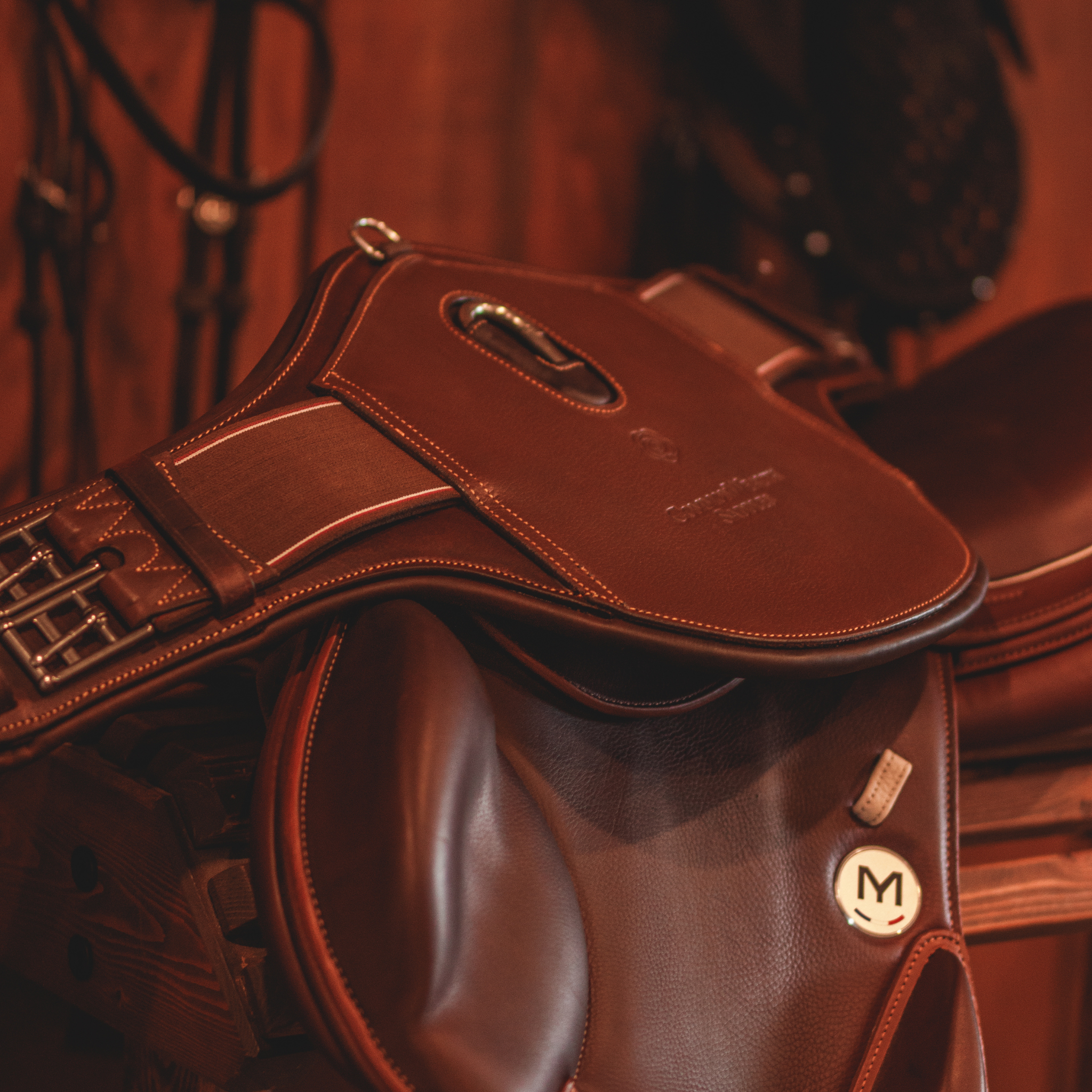 A brown leather short stud girth draped over a Meyer monoflap saddle set against a backdrop of equestrian gear in a warmly lit tack room.