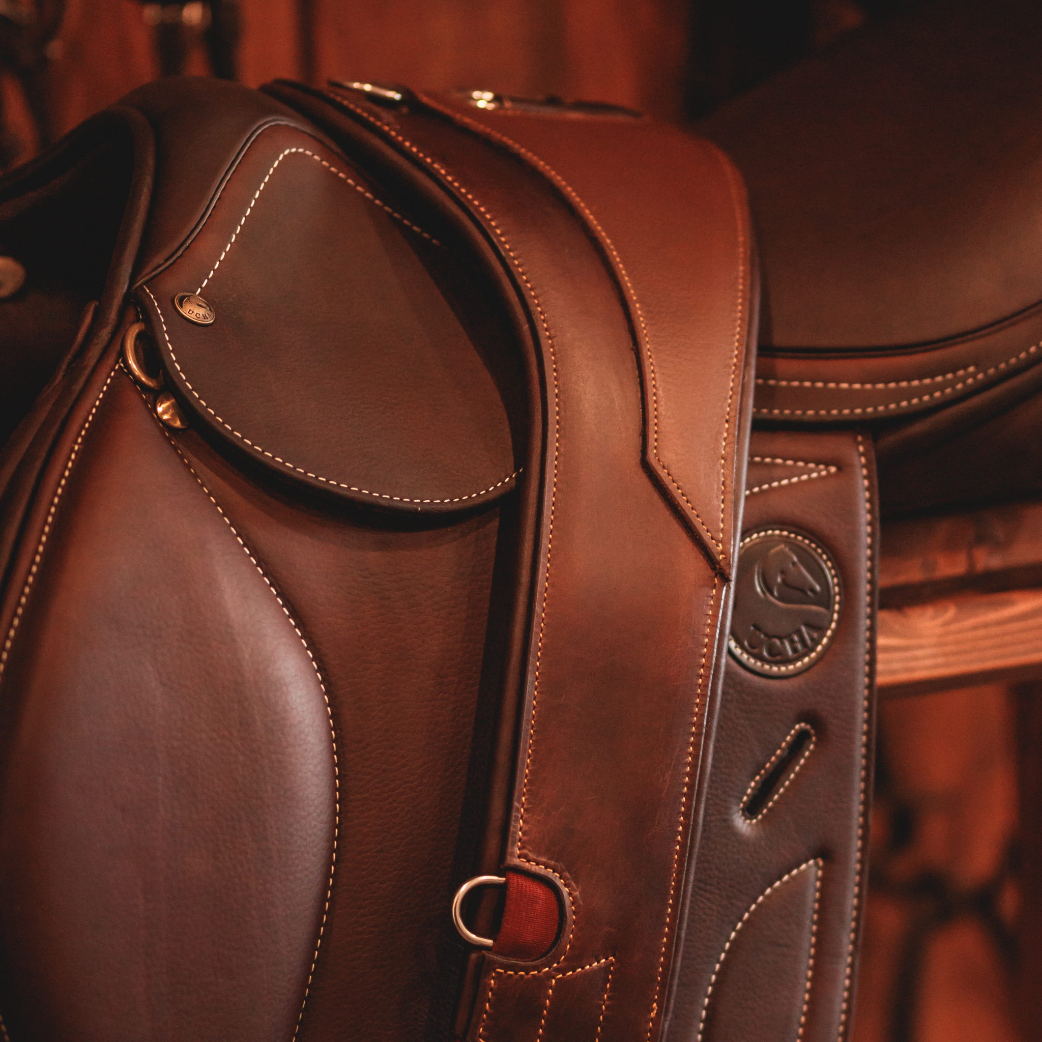 Close-up of a brown leather anatomical girth with d-ring attachments, draped over a brown Ucha jumping saddle. 