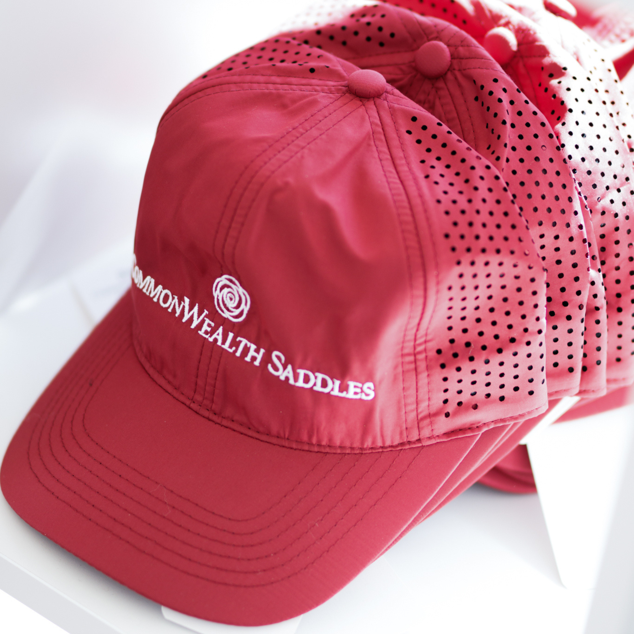 A bright burgundy ball cap with the logo 'CommonWealth Saddles' on the front and a laser perforated back.