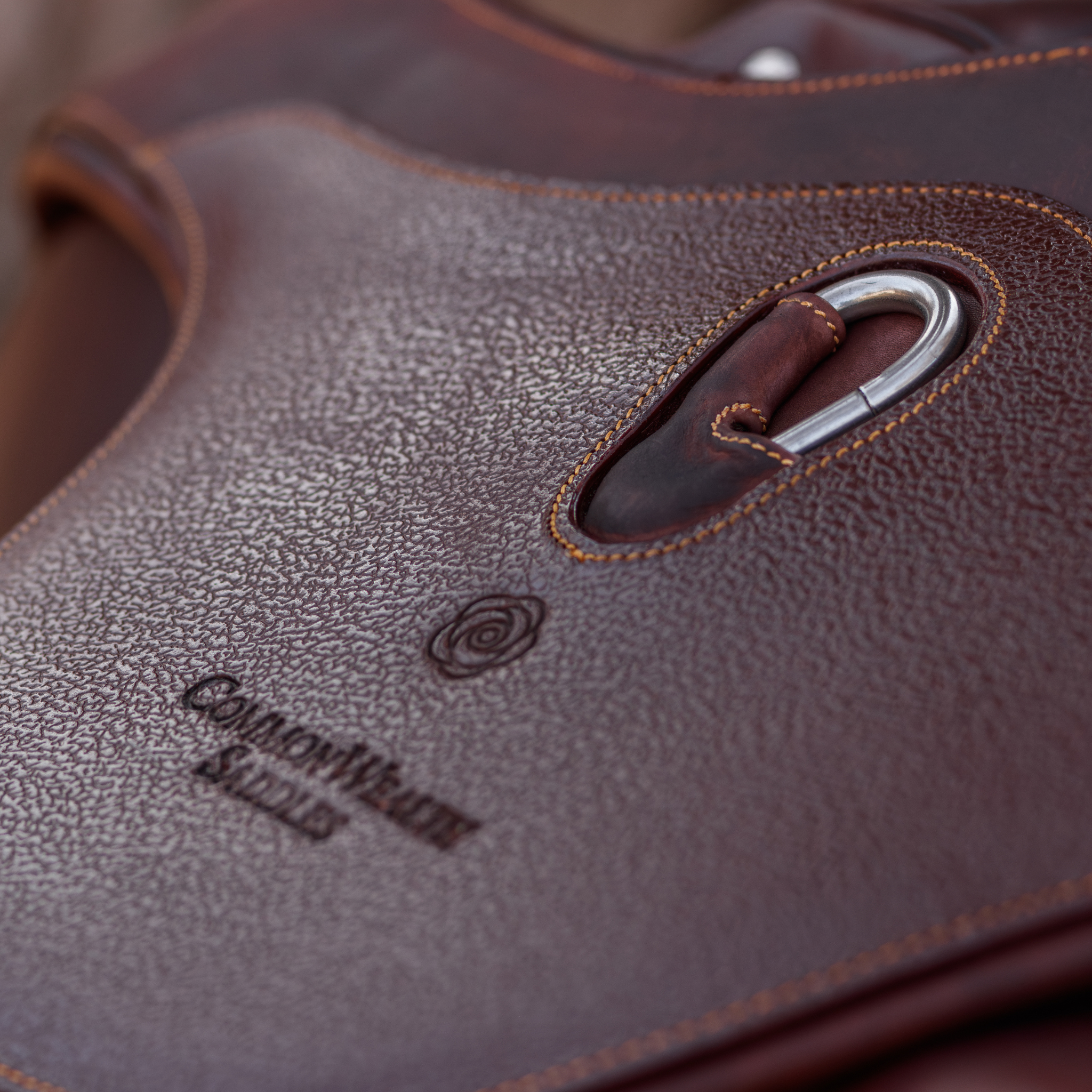 Close-up image of the centre belly pad of a brown leather Hudson Long Anatomical Stud Girth. The girth has a grain leather overlay, centre carabiner, caramel stitching, and "CommonWealth Saddles" logo hot pressed into the leather. 