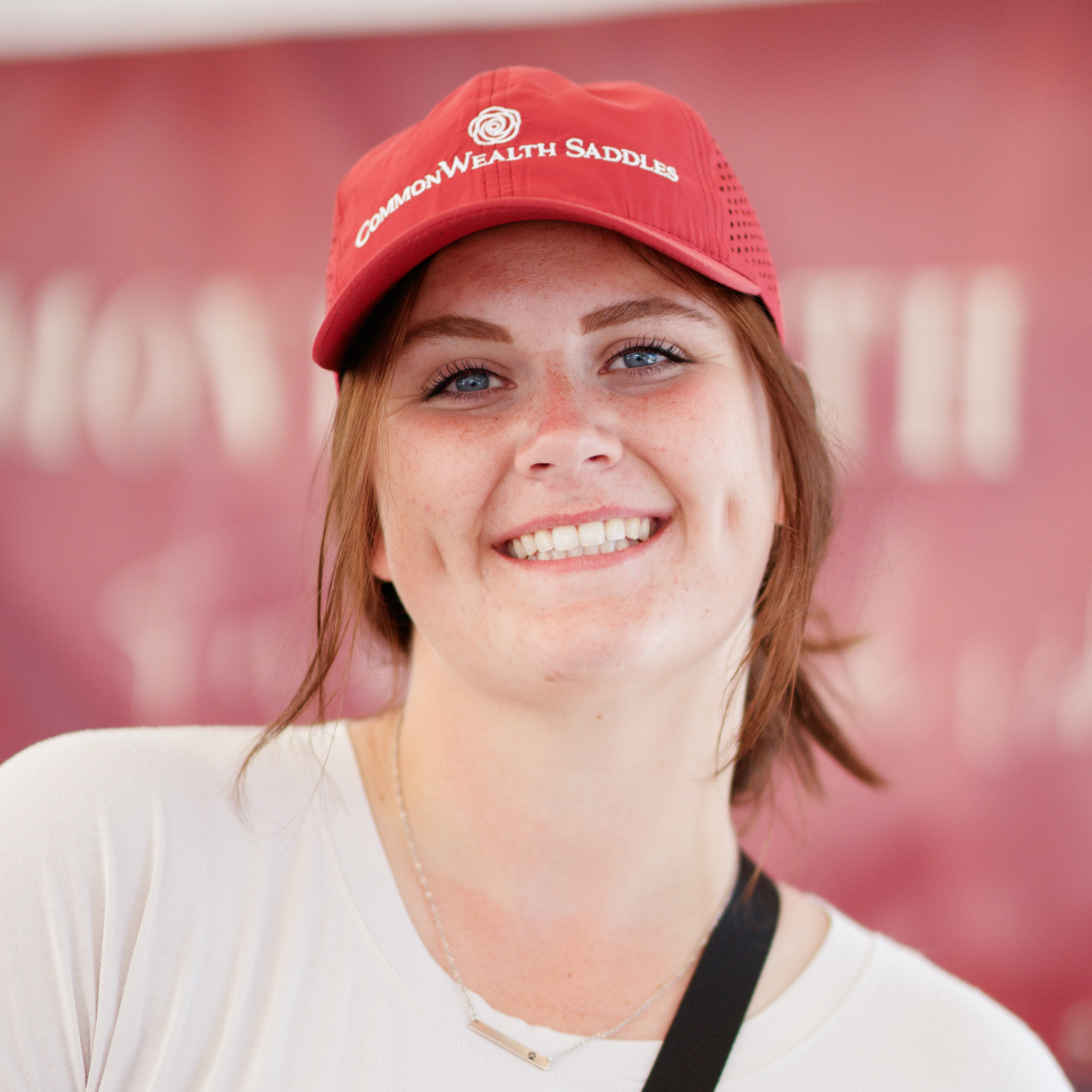 Photo of a smiling woman in a bright burgundy ball cap with the logo 'CommonWealth Saddles' on the front and a laser perforated back.