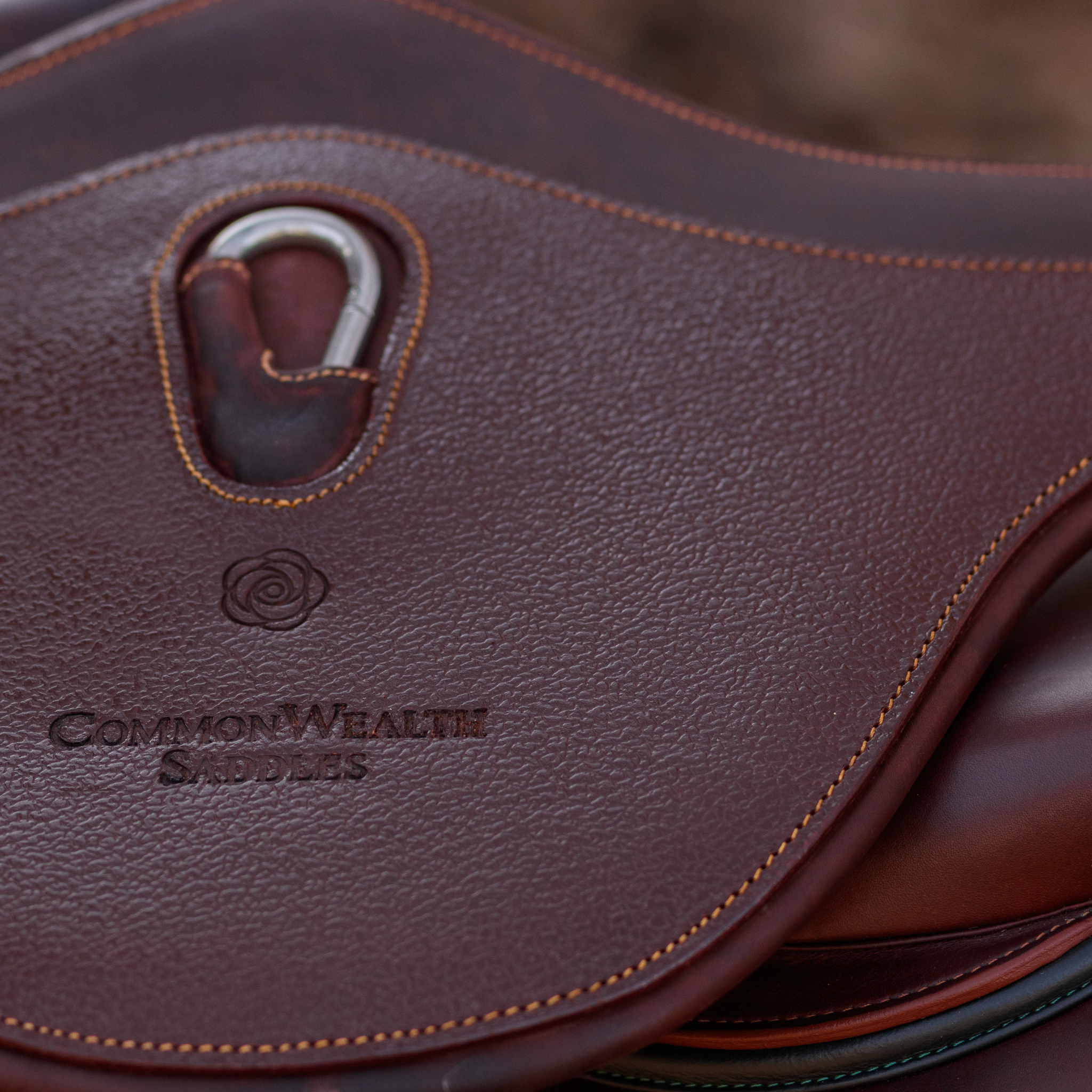 Close-up image of the centre belly pad of a brown leather Hudson Long Anatomical Stud Girth. The girth has a grain leather overlay, centre carabiner, caramel stitching, and "CommonWealth Saddles" logo hot pressed into the leather. 
