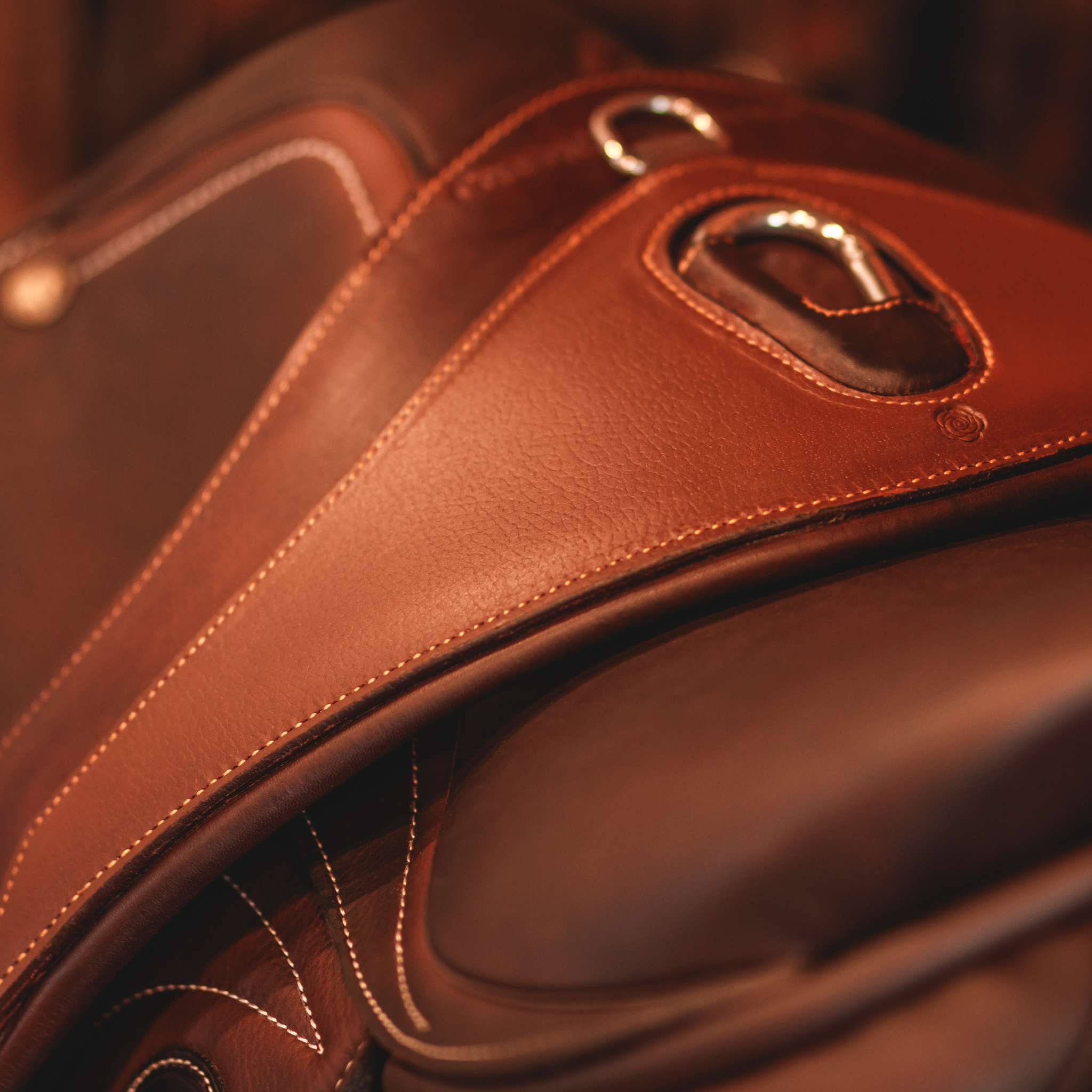 Close-up of a brown leather anatomical girth with d-ring and carabiner attachments, draped over a brown Ucha jumping saddle. 