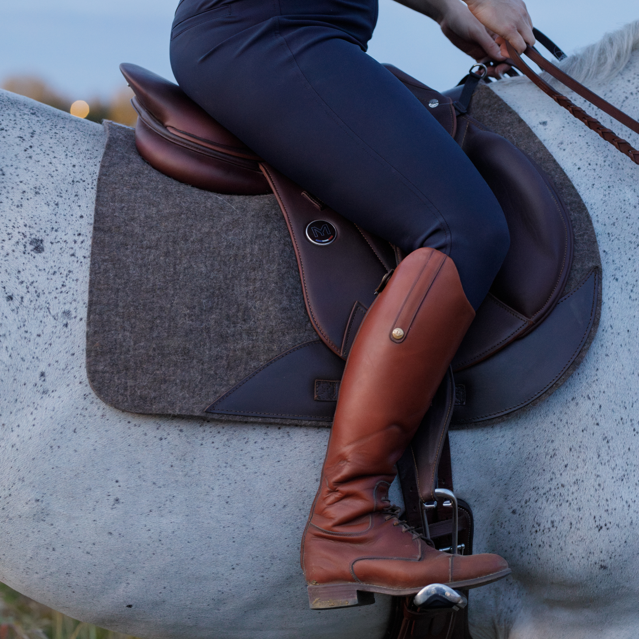 Person riding a grey horse in a Meyer saddle and natural felt half pad, with Ophena stirrups and brown Cavell leathers.