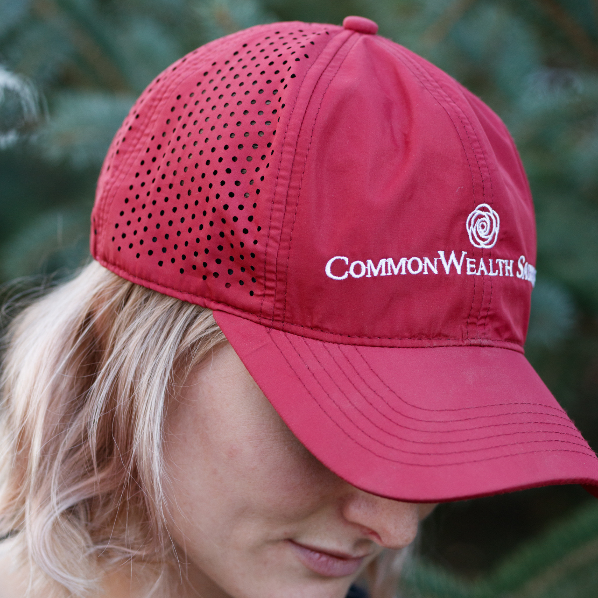 Photo of a woman wearing a bright burgundy ball cap with the logo 'CommonWealth Saddles' on the front and a laser perforated back.
