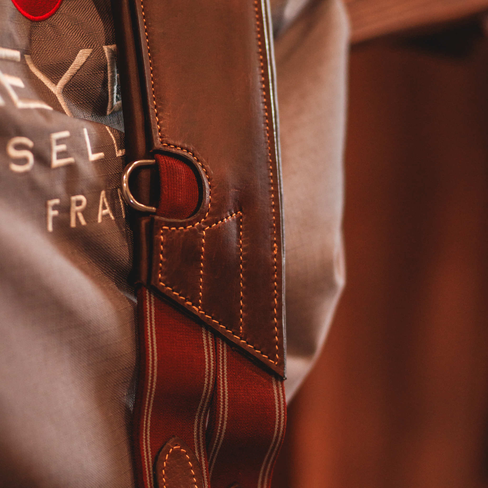 Close-up of a brown leather anatomical girth with d-ring attachment and burgundy elastics, draped over a saddle in a grey Meyer saddle cover. 