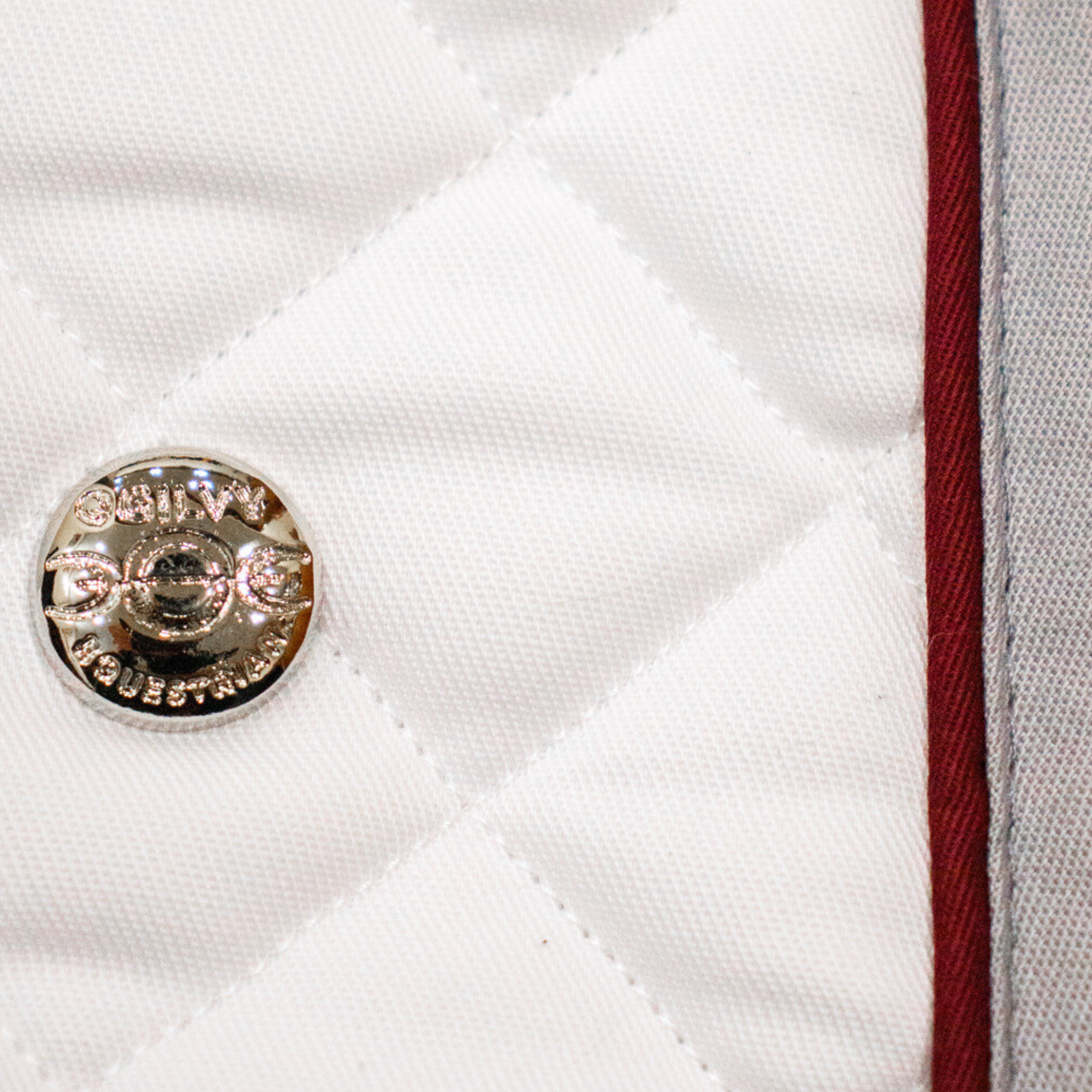 Close up of a quilted white Ogilvy saddle pad with grey binding and burgundy piping, and a metal ogilvy logo.