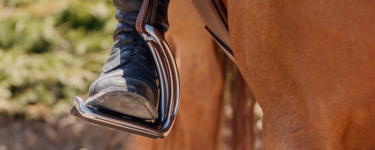 Close-up of a rider's foot in an Ophena magnetic stirrup and brown stirrup leather while mounted on a chestnut horse. 
