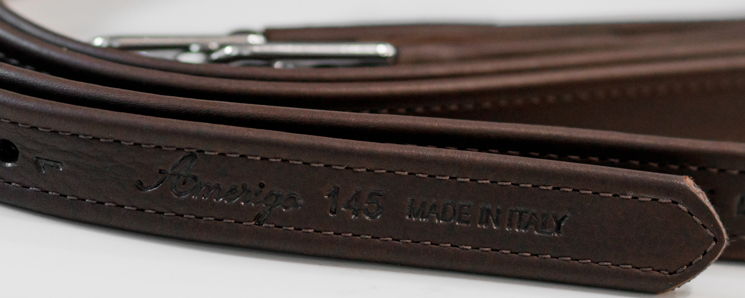 Close-up of a dark brown stirrup leather with a silver metal buckle, featuring a hot pressed "Amerigo" logo and 'MADE IN ITALY' text on the tail end.