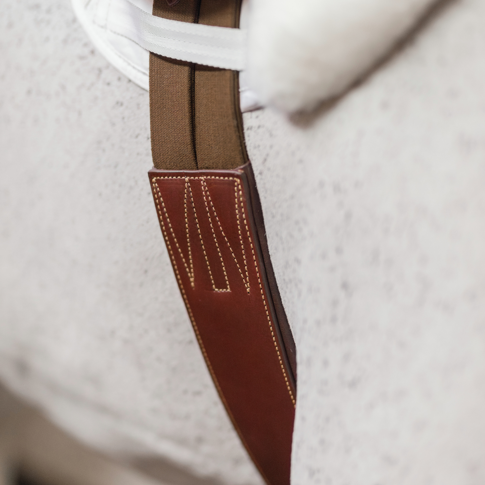 Close-up of a brown leather hunter girth on a grey horse. The girth has brown elastics and caramel stitching.