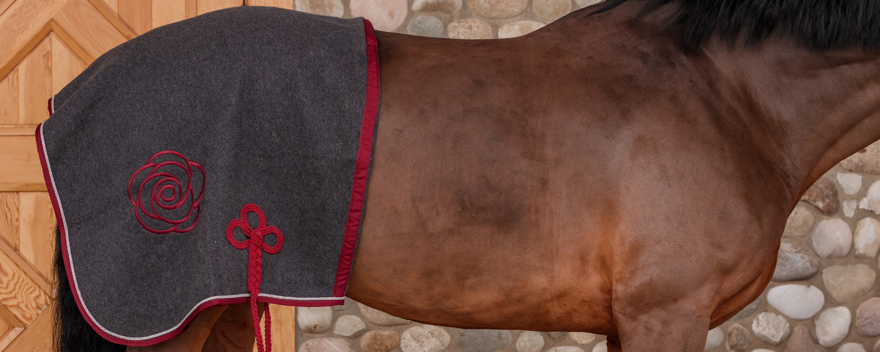 Bay horse wearing a gray rug with a burgundy peony design on a stone wall background.