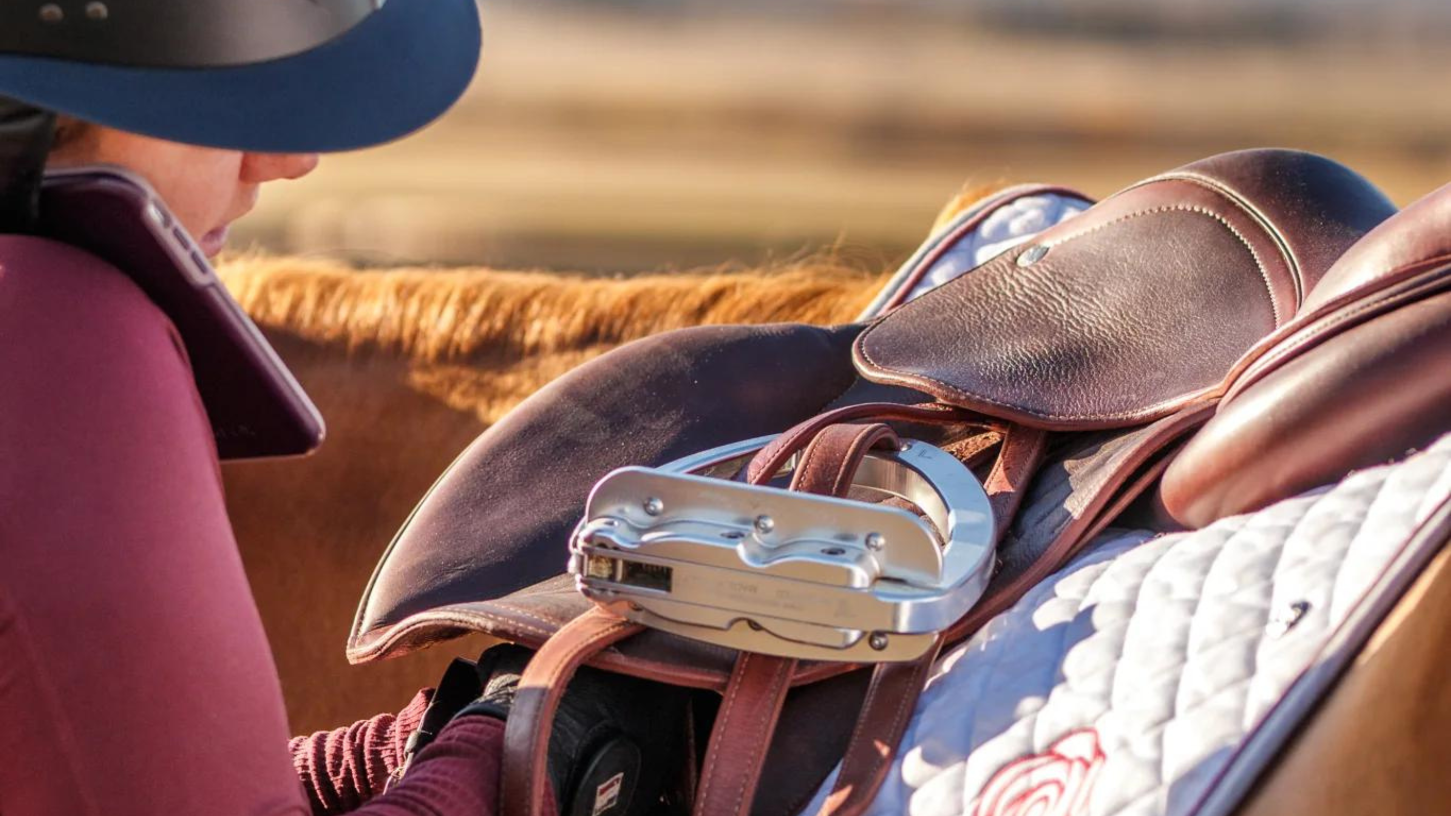 Close up photo of a rider standing next to a horse, doing up the girth while talking on the phone at the same time