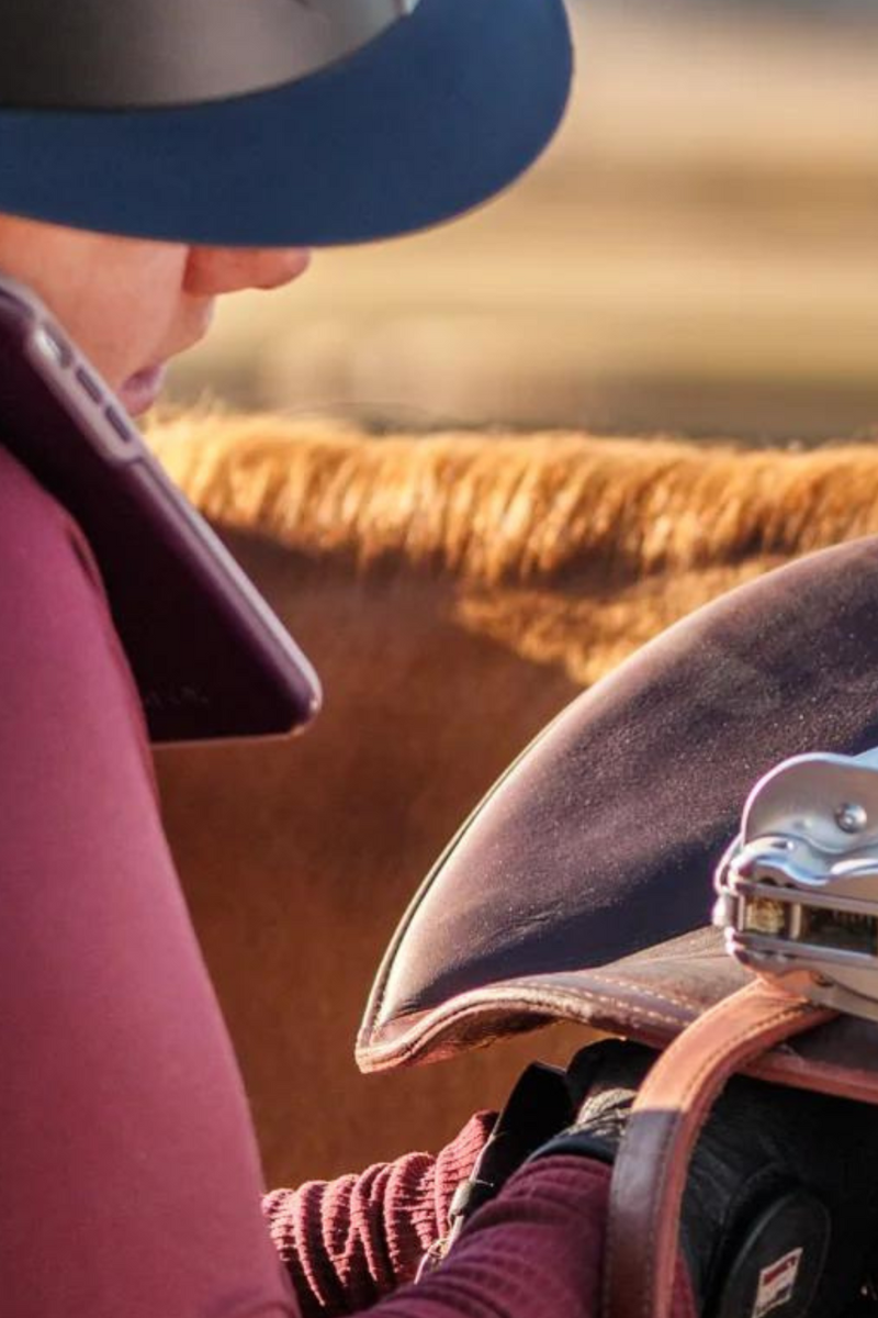Close up photo of a rider standing next to a horse, doing up the girth while talking on the phone at the same time