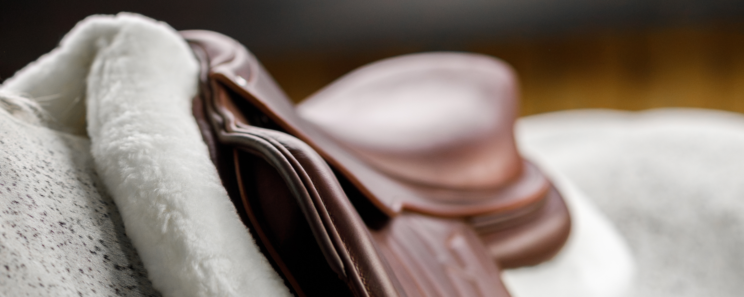 A close-up of a grey horse wearing a brown leather saddle with a a fluffy white Leveza shaped hunter pad underneath.