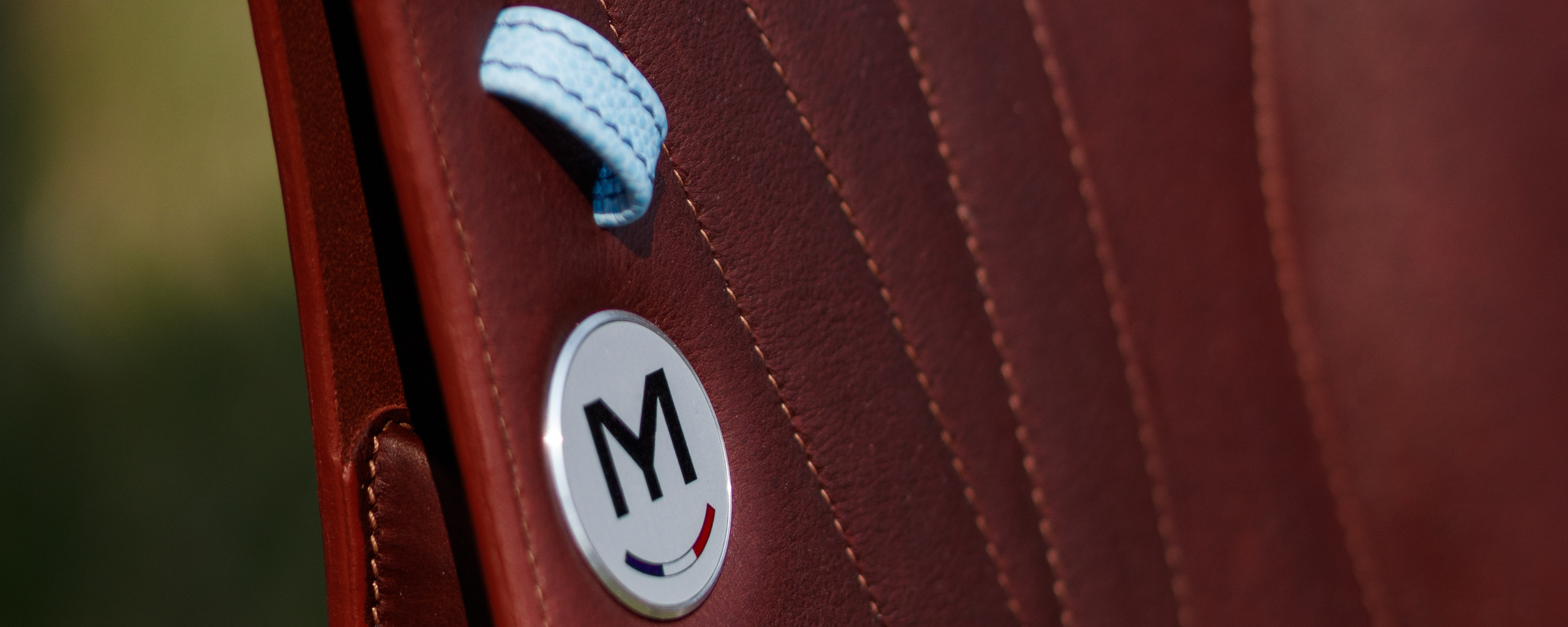 Close-up of a brown leather jump saddle with a light blue stirrup leather keeper, and a circular white badge featuring a black letter 'M' and a small French tricolor detail.