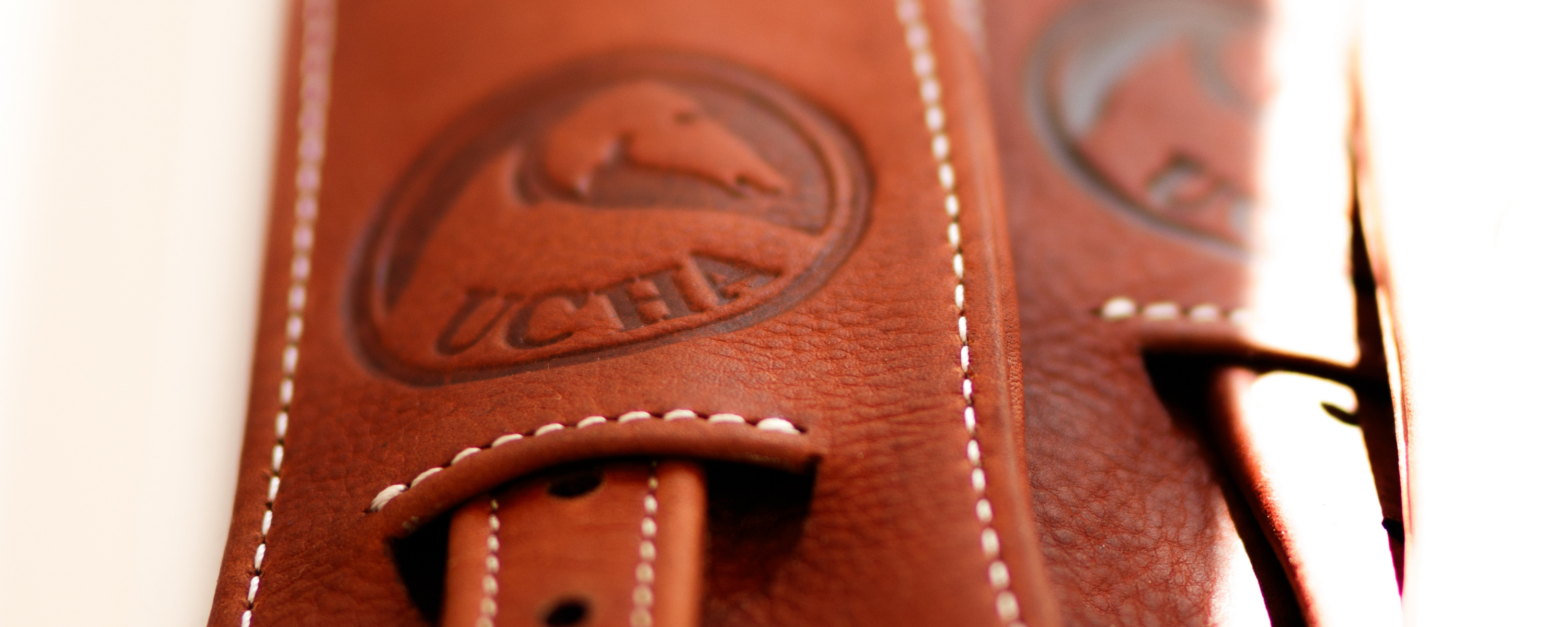 Close-up of a brown Ucha wide stability stirrup leather with white stitching and an embossed circular logo featuring a horse head and the text 'UCHA'.