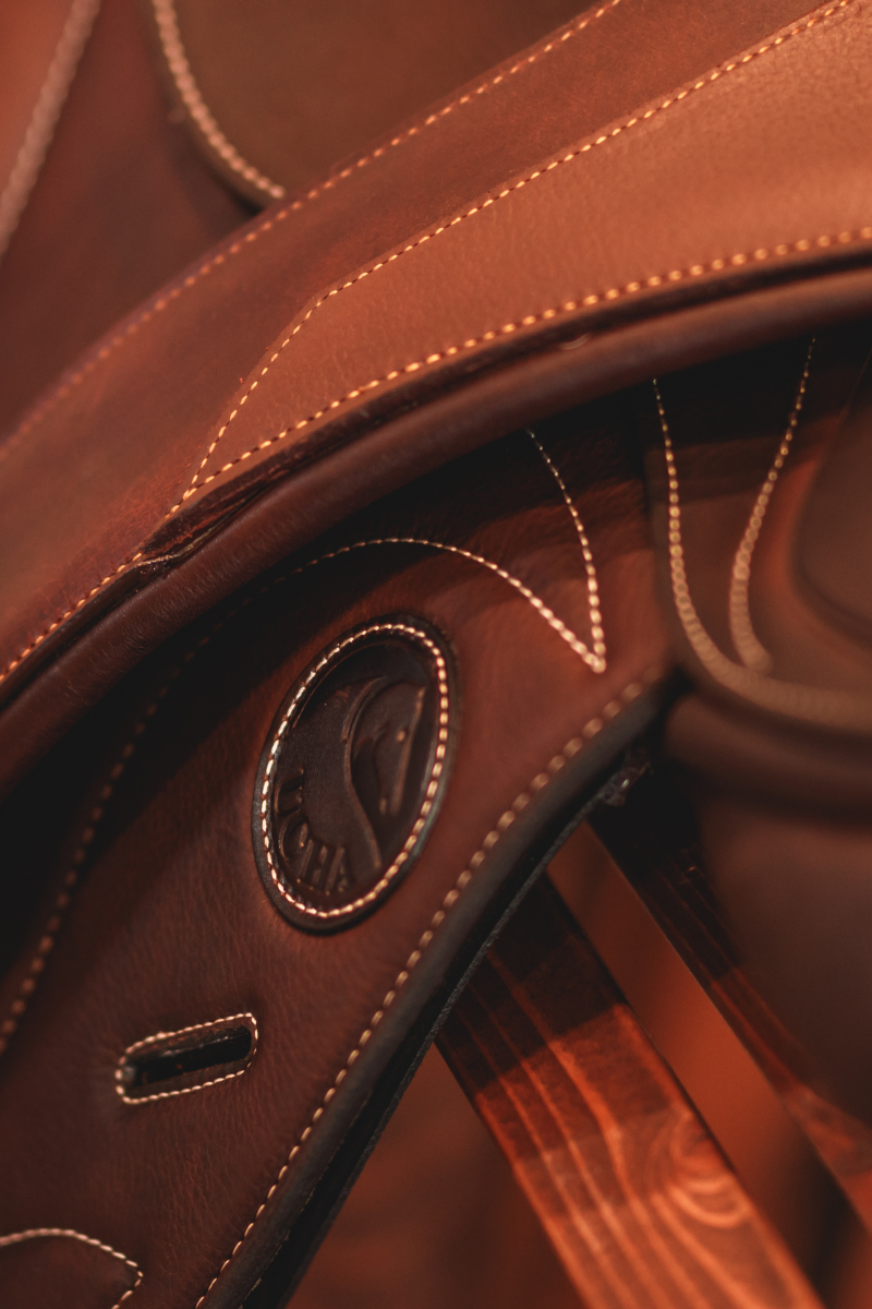 Close up photo of a rich brown Ucha saddle with a CommonWealth Rideau girth draped over top.