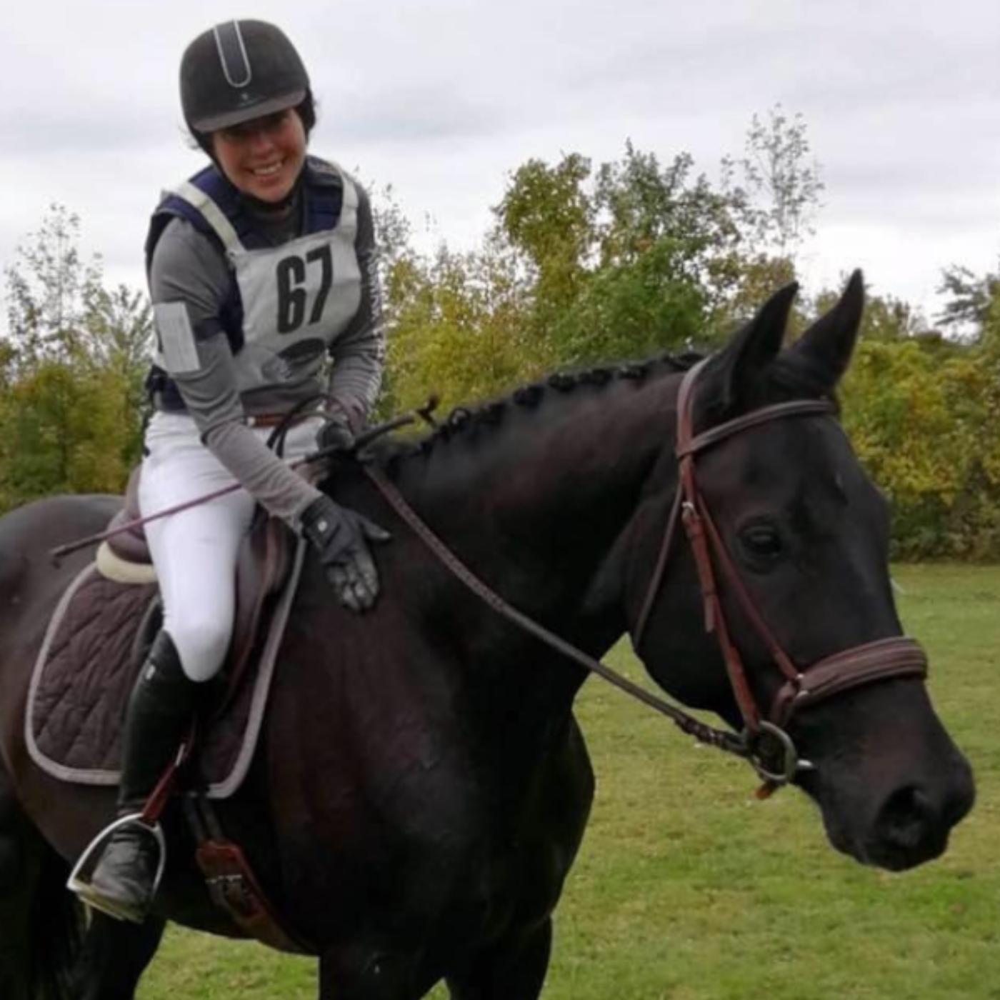 Woman riding a black horse in equestrian attire with a number 67 on a grassy field.