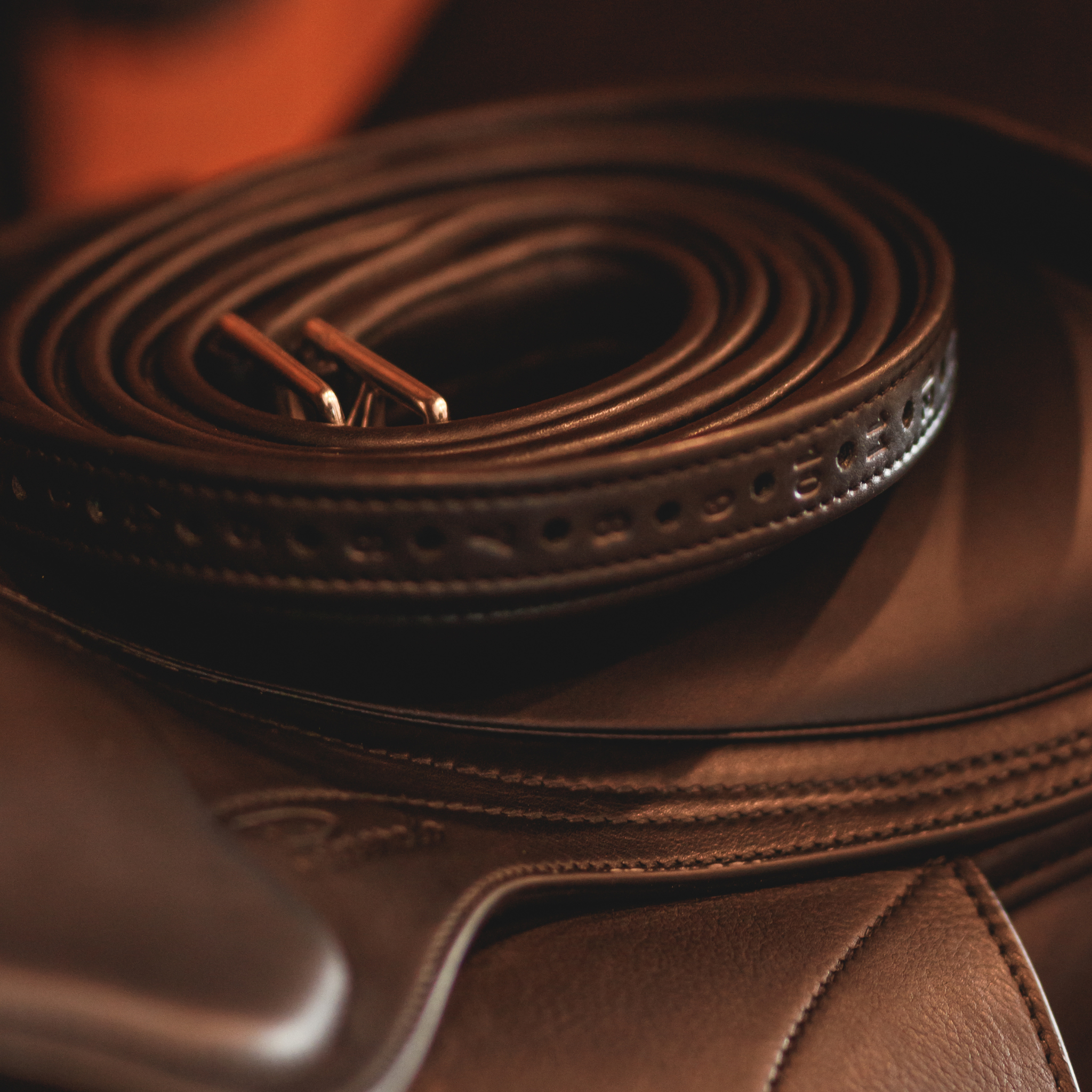 Black Yoho rolled stirrup leathers sitting on top of a black dressage saddle against a blurred brown background. 